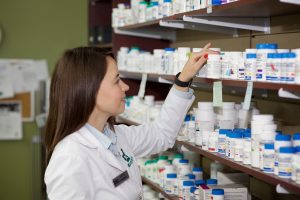 Female pharmacist in lab coat pulling medication bottle from shelf.